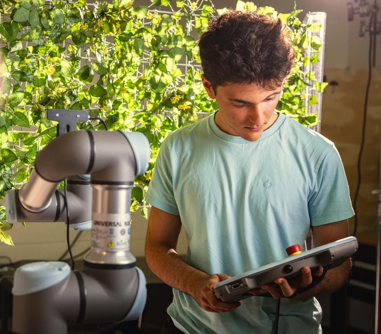 Holding the teach pendant beside the Universal Robots arm, in front of the lab's flower wall